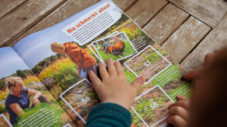 1E9A9858_stickeralbum Kinderhände blättern in einem Magazin oder Buch, das Bilder und Text über Bio-Landwirtschaft zeigt. Zu sehen sind ein Junge mit Gießkanne in einem Garten, ein Hahn und Hühner sowie Textabschnitte mit Informationen zu Bio-Produkten. Der Hintergrund ist ein Holztisch.